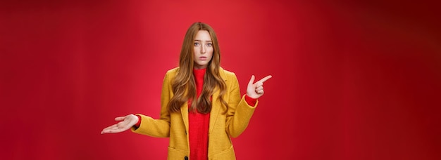 Zdjęcie sfrustrowana i zdezorientowana młoda śliczna kobieta z rudymi włosami w żółtym płaszczu, wzruszająca ramionami jako wskazująca na pytania