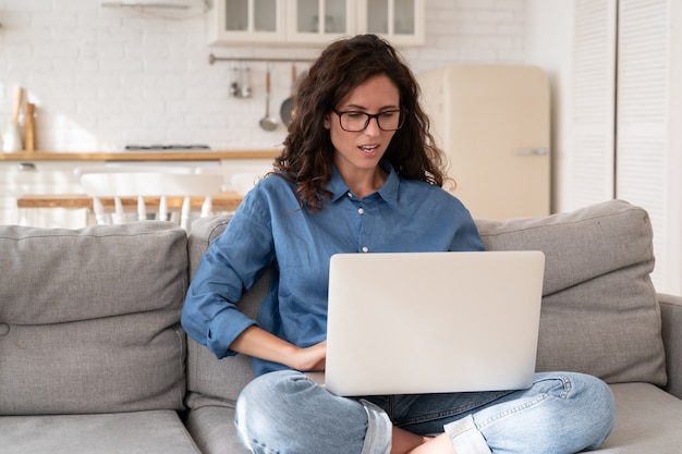 Sfrustrowana kobieta patrzy na ekran laptopa z otwartymi ustami i nieufnością na twarzy, siedząc na kanapie w domu
