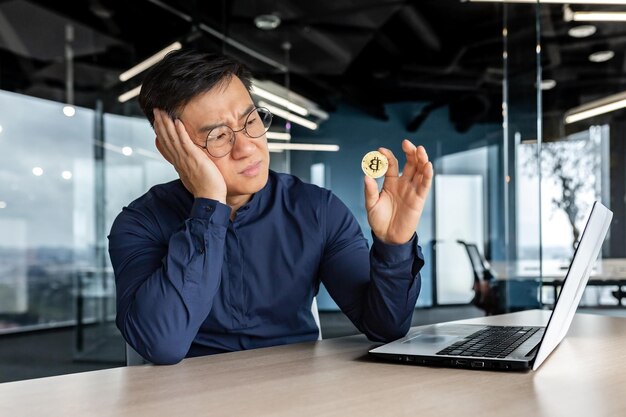 Sfrustrowany azjatycki biznesmen inwestor trzymający kryptowalutę bitcoin w rękach mężczyzna pracujący w środku
