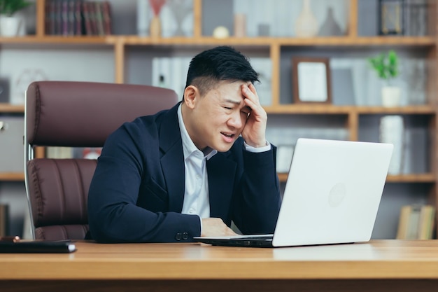 Sfrustrowany i zły azjatycki biznesmen pracujący na laptopie w biurze szefa krzyczącego na ekranie laptopa