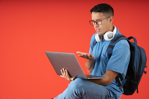 Sfrustrowany młody człowiek pracujący na laptopie w słuchawkach i niosący plecak