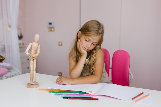 Siedmioletnia uczennica wykonuje zadania domowe przy biurku w domu lub w szkole, rysuje długopisem i zastanawia się nad notatnikiem