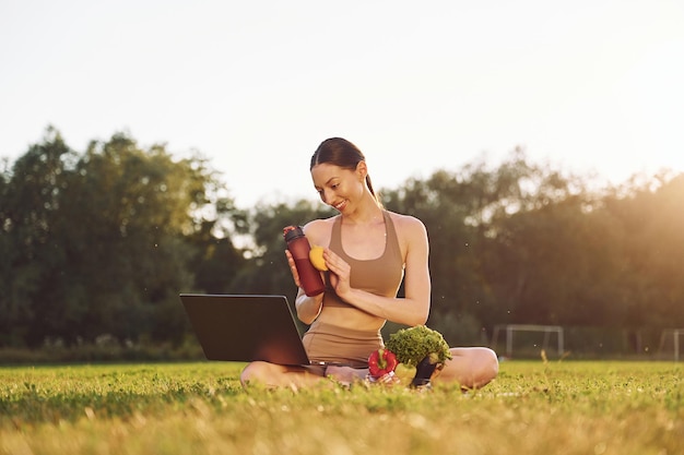 Siedząc z laptopem i warzywami Młoda kobieta w ubraniach jogi jest na zewnątrz na polu