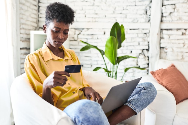 Siedząca na kanapie afrykańska kobieta robi zakupy w Internecie
