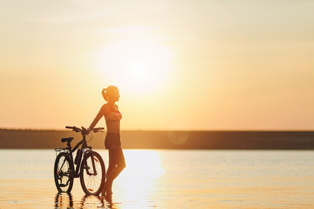 Silna blondynka w kolorowym garniturze stoi obok roweru w wodzie o zachodzie słońca w ciepły letni dzień. Koncepcja fitness.