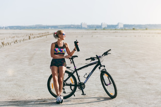 Silna Blondynka W Wielobarwnym Garniturze I Okularach Przeciwsłonecznych Stoi Przy Rowerze Z Czarną Butelką Wody Na Pustyni I Patrzy Na Słońce. Koncepcja Fitness.