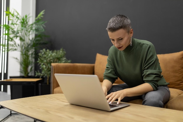 Silna kobieta z krótką fryzurą pracująca na laptopie siedząc na kanapie w domu