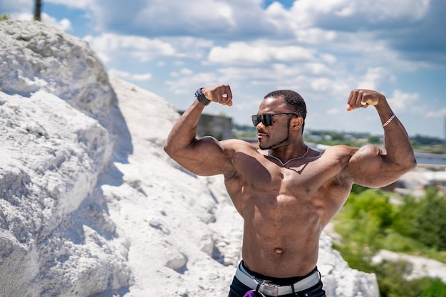 Silnie wysportowany kulturysta pokazujący bicepsy Przystojny wysportowany mężczyzna pokazujący swoje mięśnie stojąc na piasku z błękitnym niebem w tle