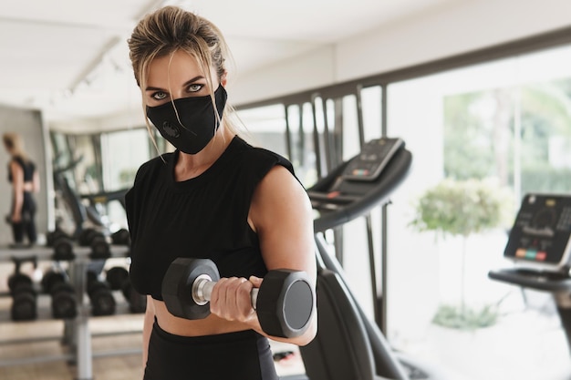 Siłownia nowa normalna. Młoda kobieta lekkoatletycznego noszenie maski profilaktycznej podczas treningu fitness z hantlami.