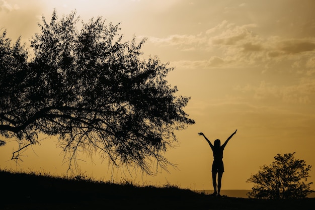 Silueta szczęśliwej kobiety tańczącej o zachodzie słońca obok dużego drzewa z rękami w górę do nieba
