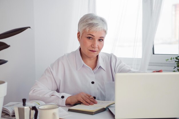 Siwowłosa bizneswoman pracuje przy biurku z komputerem patrząc na kamerę