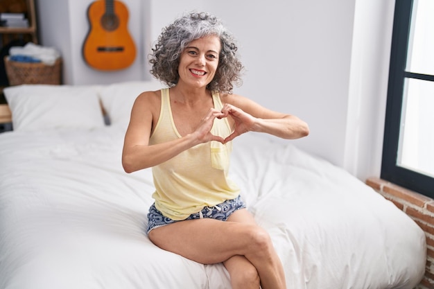 Siwowłosa kobieta w średnim wieku robi gest serca siedząc na łóżku w sypialni