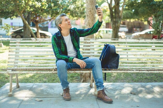 Siwowłosy mężczyzna siedzi na ławce w parku i robi sobie selfie