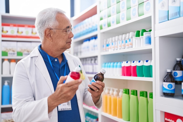 Siwowłosy mężczyzna w średnim wieku, farmaceuta, uśmiechający się pewnie, trzymając butelki z lekami w aptece