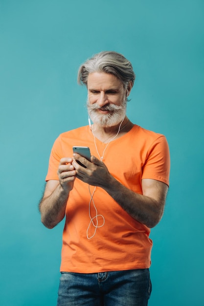 Siwy starszy mężczyzna w przewodowych słuchawkach z telefonem pozuje na niebieskim tle Strzelanie w studio