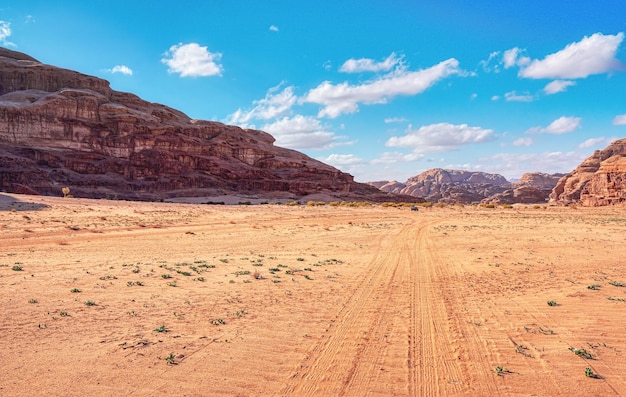 Skaliste masywy na czerwonej pustyni piaskowej, jasne pochmurne niebo w tle, mały pojazd z napędem na 4 koła i wielbłąd na odległość - typowa sceneria w Wadi Rum, Jordania