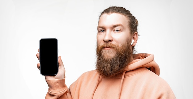 Skandynawski Przystojny Poważny Mężczyzna Trzymający Telefon Z Makietą Czarnego Ekranu Z Bezprzewodowymi Słuchawkami Na Białym Tle Na Szarym Tle Spokojny Facet Z Rudą Fryzurą I Brodą Nowoczesne Technologie Cyfrowe