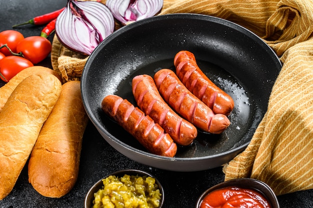 Składniki Do Robienia Domowych Hot-dogów. Kiełbaski Na Patelni, świeże Upieczone Bułki, Musztarda, Keczup, Ogórki. Czarne Tło. Widok Z Góry