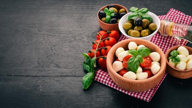 Składniki na włoską sałatkę caprese ser mozzarella pomidorki koktajlowe liście bazylii oliwki oliwa pieprz na czarnym drewnianym tle wolne miejsce na tekst