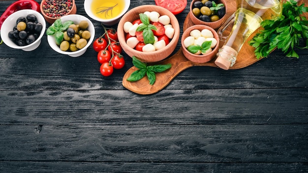 Składniki na włoską sałatkę caprese ser mozzarella pomidorki koktajlowe liście bazylii oliwki oliwa pieprz na czarnym drewnianym tle wolne miejsce na tekst