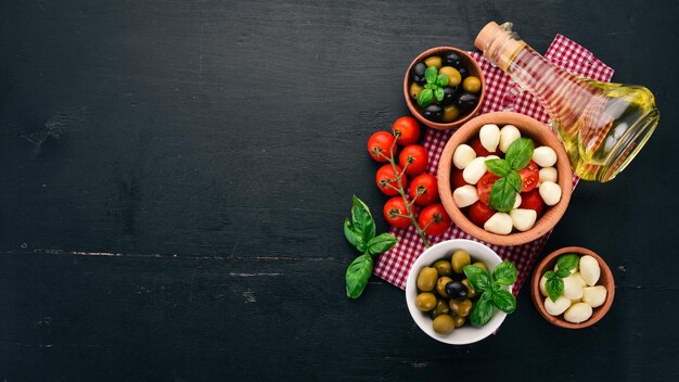 Składniki na włoską sałatkę caprese ser mozzarella pomidorki koktajlowe liście bazylii oliwki oliwa pieprz na czarnym drewnianym tle wolne miejsce na tekst
