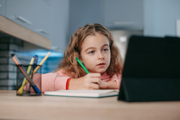 Skoncentrowana dziewczyna słucha lekcji online, patrząc na ekran tabletu i robiąc notatki online, edukator