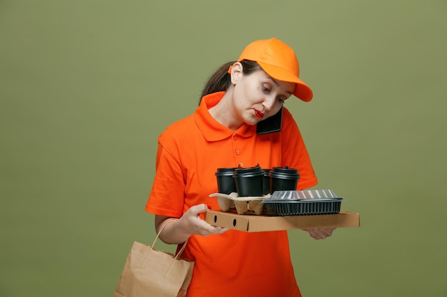 Skoncentrowana Kobieta Dostarczająca W średnim Wieku Ubrana W Mundur I Czapkę Trzymająca Opakowanie Pizzy Pojemnik Na Jedzenie Taca Na Jajka Plastikowe Kubki Do Kawy I Papierową Torbę Patrzącą W Dół Rozmawiającą Przez Telefon