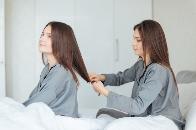 Skoncentrowana ładna młoda kobieta w szarej piżamie siedząca na łóżku i czesząca bliźniacze włosy swojej siostry