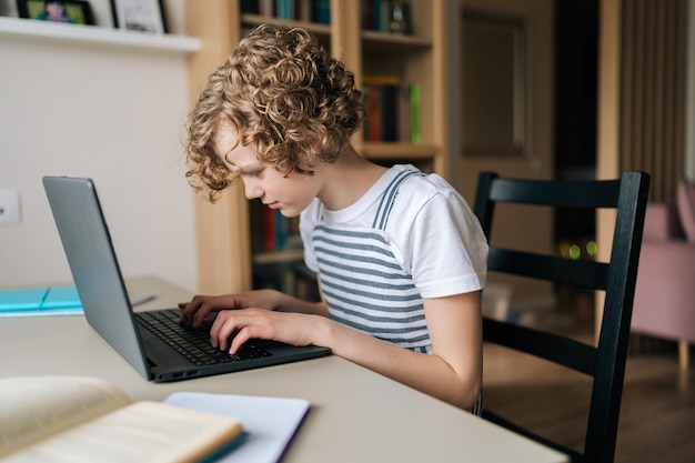 Skoncentrowana mała uczennica pracująca nad pracą domową, wpisując informacje na laptopie, studiując online w domu, siedząc przy biurku przy oknie