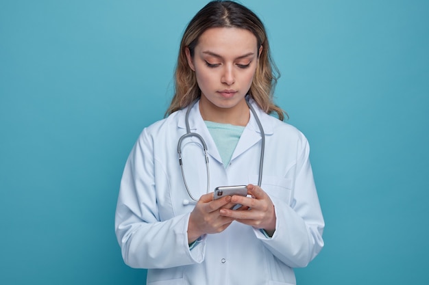Skoncentrowana młoda kobieta lekarz ubrana w szlafrok i stetoskop wokół szyi przy użyciu swojego telefonu komórkowego