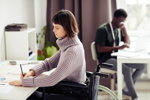 Skoncentrowana młoda krawcowa siedząca na wózku inwalidzkim przy biurku i poprawiająca wykrój krawiecki dla klienta