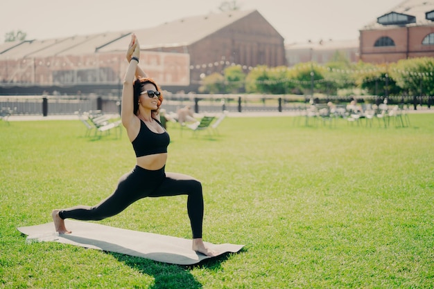 Skoncentrowana pozytywna kobieta podnosi ramiona, wykonuje ćwiczenia pilates, ubrana w okulary przeciwsłoneczne, ma trening podczas letnich praktyk jogi na zielonej trawie Koncepcja ludzi i zdrowego stylu życia