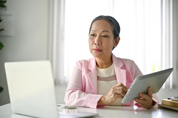 Skoncentrowana starsza azjatycka bizneswoman używająca laptopa i tabletu przy biurku w biurze