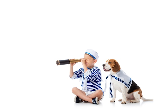 Skoncentrowane dziecko w wieku przedszkolnym w stroju marynarza patrzące w lunetę siedząc z psem rasy beagle