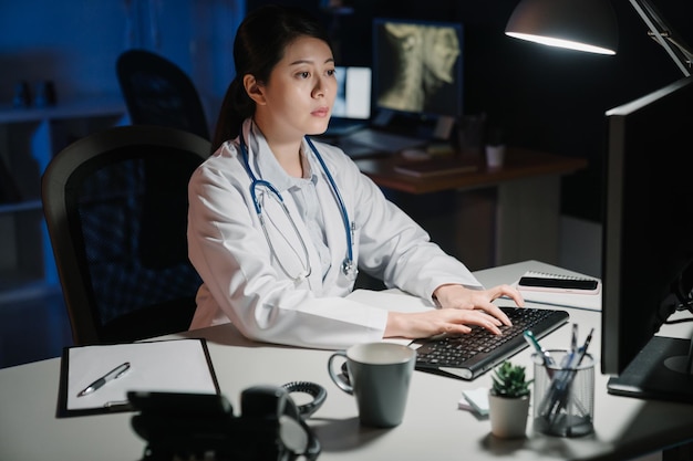 Skoncentrowany azjatycki chiński lekarka z raportem patrząc na monitor komputera przy biurku w gabinecie lekarskim w nocy. młoda kobieta personel kliniki późno spać w miejscu pracy w szpitalu i pisania.