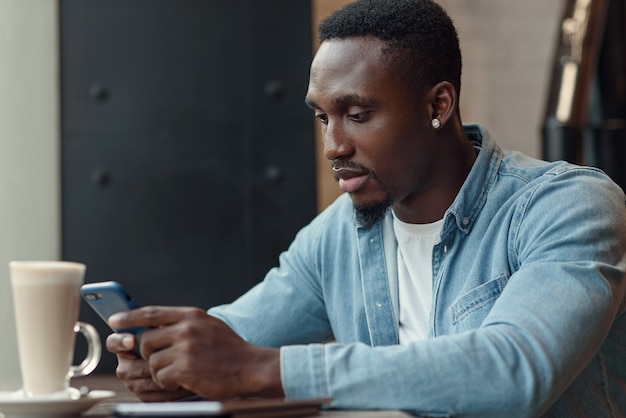 Skoncentrowany czarny mężczyzna przedsiębiorca używa smartfona siedząc w kawiarni z kawą przy oknie