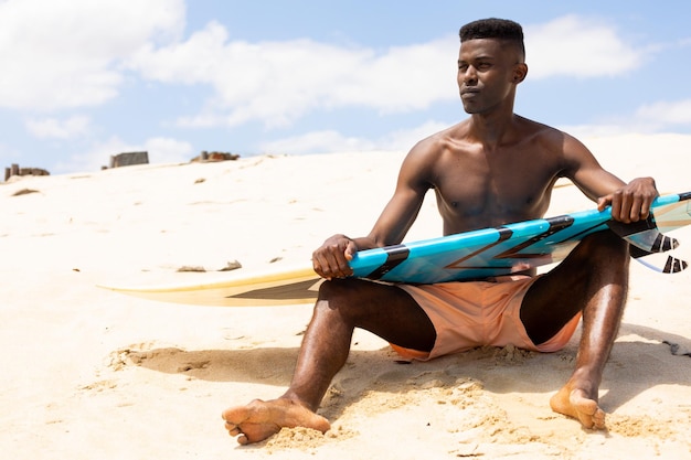 Skoncentrowany dwurasowy mężczyzna trzymający deskę surfingową siedzący na słonecznej plaży i odwracający wzrok. Aktywny tryb życia, hobby, sport, surfing, lato i wakacje, bez zmian.