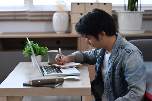 Skoncentrowany freelancer pracujący online z laptopem i robiący notatki na notebooku