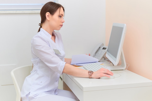 Skoncentrowany lekarka z raportowym patrzeje komputerowym monitorem przy biurkiem w medycznym biurze.