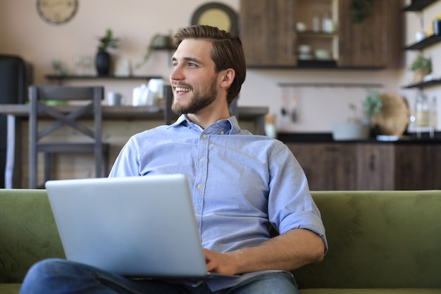 Skoncentrowany młody biznesmen freelancer siedzący na kanapie z laptopem, pracujący zdalnie online w domu.