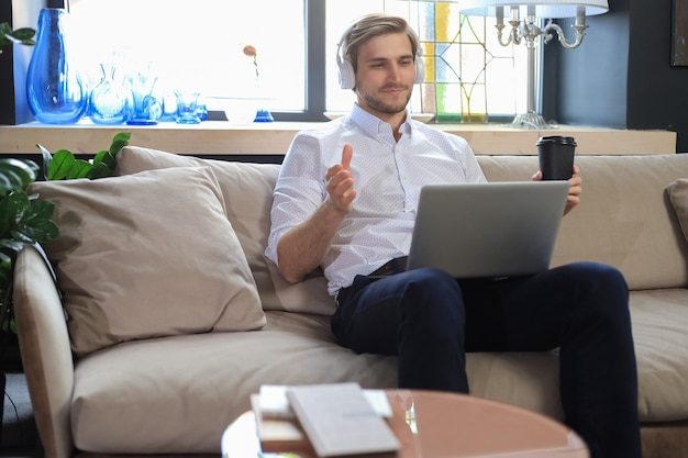 Skoncentrowany młody biznesmen freelancer siedzący na kanapie z laptopem, pracujący zdalnie online w domu.