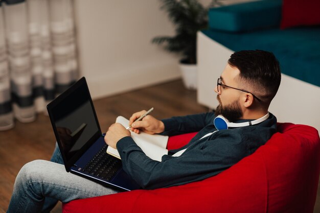 Skoncentrowany młody człowiek nosi słuchawki i okulary studiuje online oglądając seminarium internetowe na laptopie. Robi notatki w zeszycie.