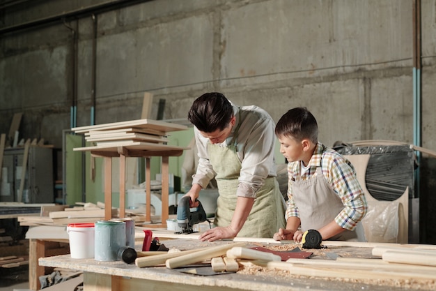 Skoncentrowany stolarz w średnim wieku w fartuchu na maszynie do polerowania pokazujący synowi, jak szlifować deski w warsztacie
