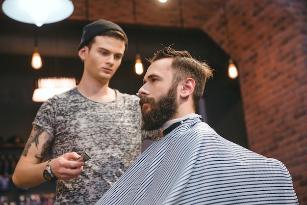 Skoncentrowany Zręczny Fryzjer Robi Fryzurę Młodemu Brodatemu Przystojnemu Mężczyźnie W Salonie Fryzjerskim