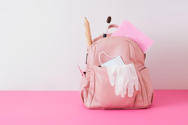 Skórzany różowy plecak na ramionach na szarym tle, maska na twarz i rękawiczki, zamknięcie w szkole