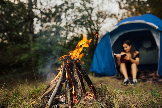 Skupić Się Na Bonfire. Młoda Kobieta Siedzi W Namiocie I Czytając Książkę W Obozie Przy Ognisku.