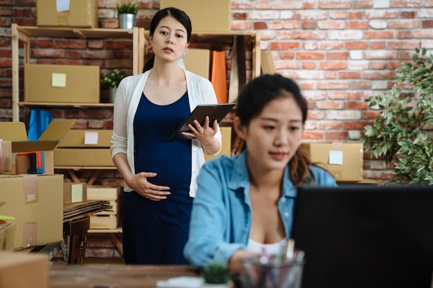 skupić się na przyszłej pracownic macierzyństwa spaceru w biurze. młoda kobieta w ciąży z cyfrowym tabletem dotykowym brzuchem działającym w małej firmie właściciel sklepu internetowego. niewyraźny widok koleżanka wpisując na komputerze przenośnym.