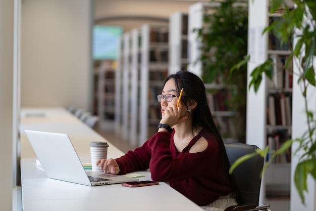 Skupiona azjatycka dziewczyna w okularach ucząca się online na laptopie w pustej bibliotece lub klasie