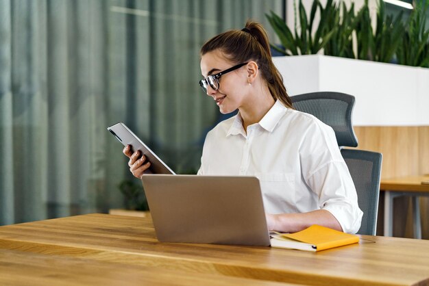 Skupiona bizneswoman porównuje dane na tabletie, jednocześnie pracując na laptopie