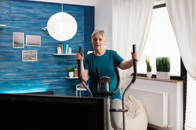 Skupiona starsza kobieta robi trening treningowy mięśni ciała oglądając wideo online ćwiczenia gimnastyczne w telewizji przy użyciu roweru wyszczuplającego w domu w salonie